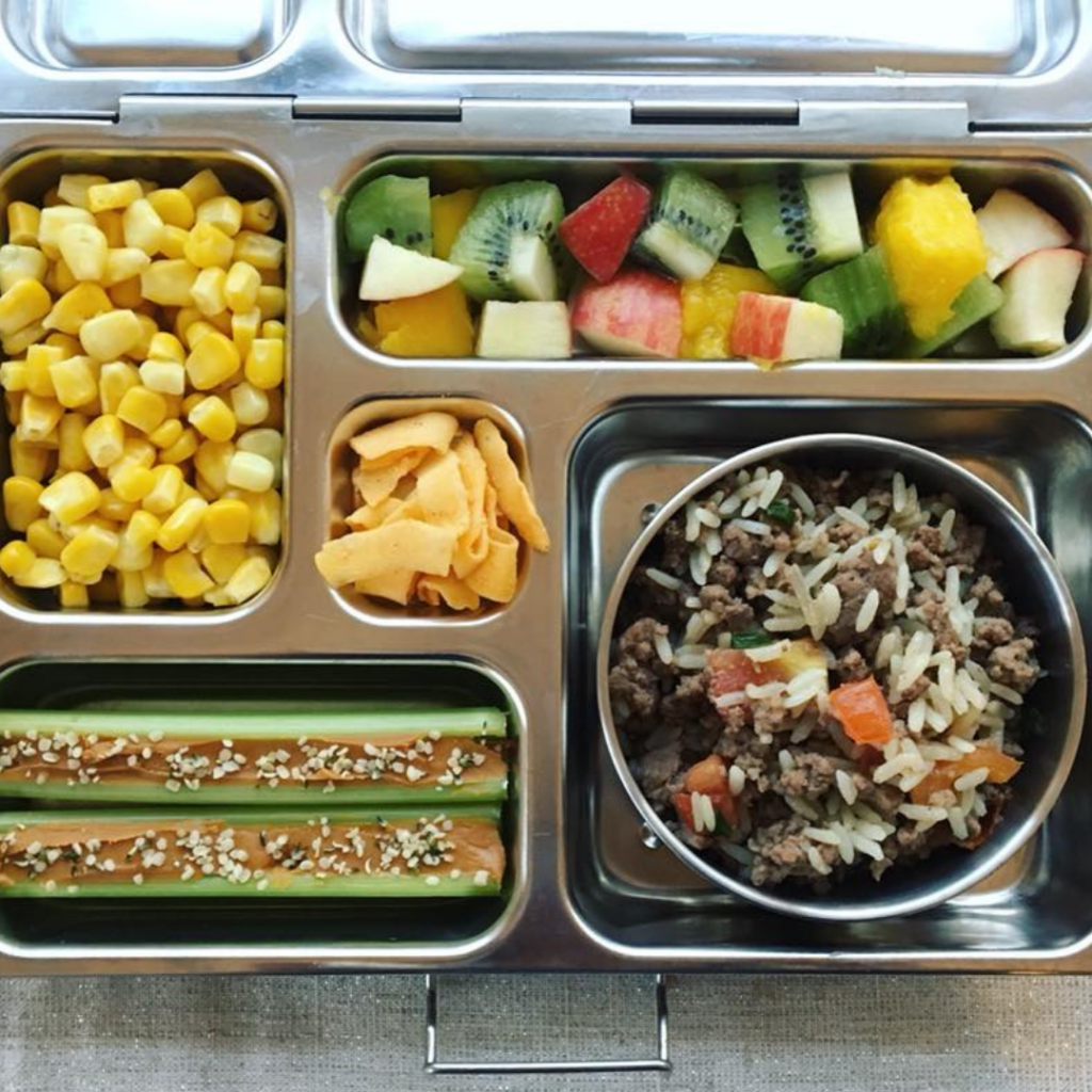 Main image for the article [The Ultimate Lunch Packing Guide]. Pictured is an open lunchbox with mixed fruit, corn, rice, hamburger and tomatoes, and celery with peanut butter on top.