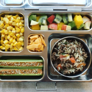  Main image for the article [The Ultimate Lunch Packing Guide]. Pictured is an open lunchbox with mixed fruit, corn, rice, hamburger and tomatoes, and celery with peanut butter on top.