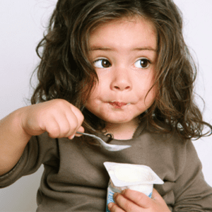 Main image for the article [The Best Yogurts For Your Baby]. Pictured is toddler eating a yogurt cup with a spoon.