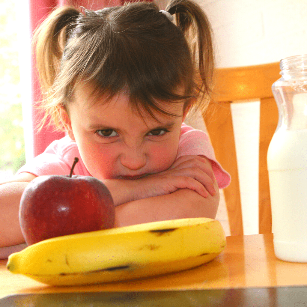 Main image for the article: "When picky eating moves from normal to extreme". Pictured is a toddler at the table looking unhappy with an apple, banana, and milk in front of them.