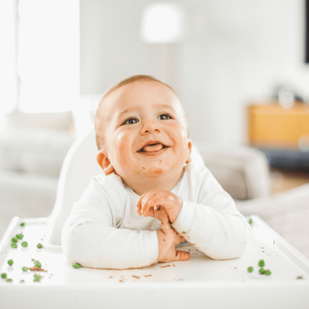 Main image for the article: "A new way to start solids with Baby Led Feeding". Pictured is a happy baby, sitting in their highchair, with peas scattered on their tray.