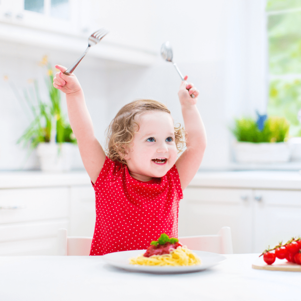 Featured image for article: "Limiting Food: Is it ever okay to control how much your toddler eats?". Pictured is a smiling toddler at a table with a plate of spaghetti in front of them.